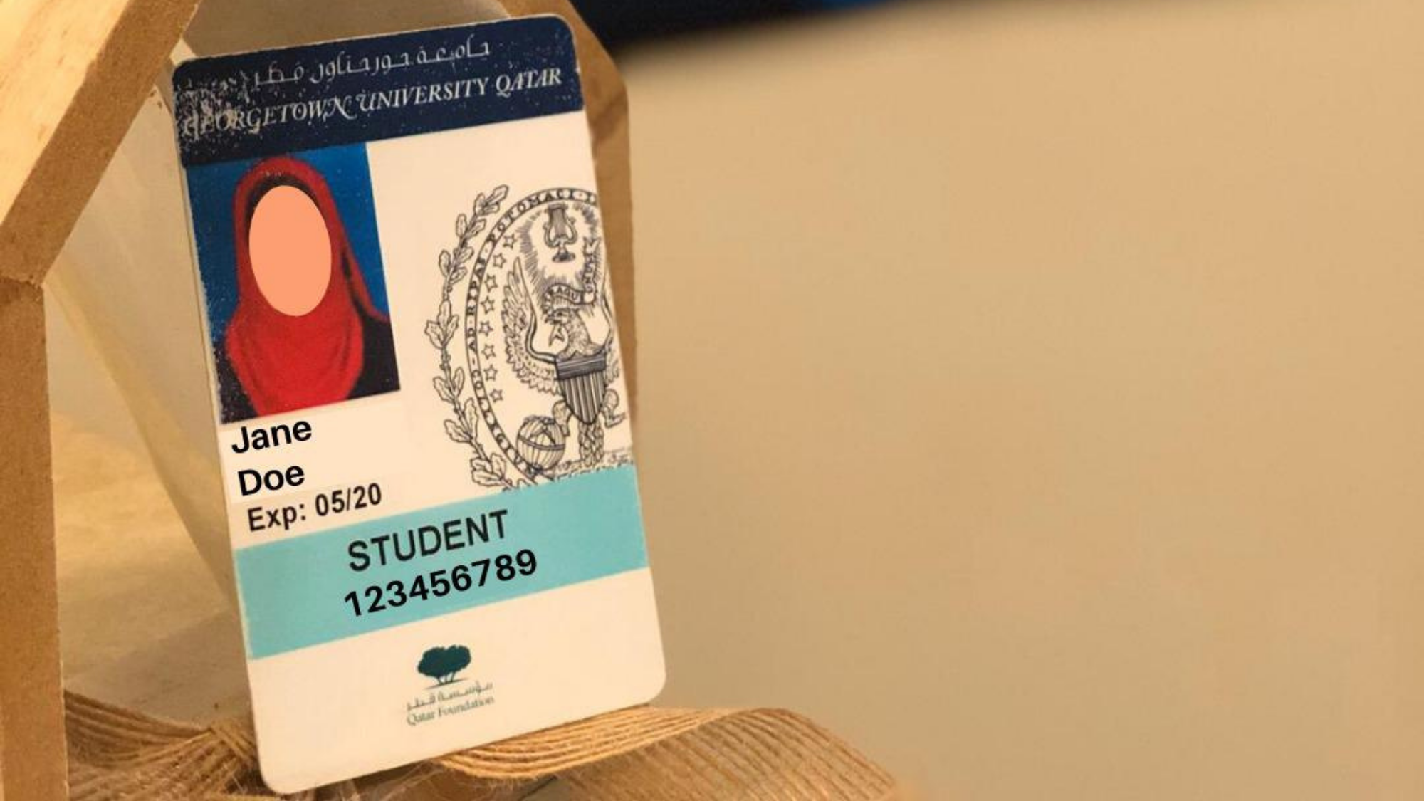 Picture of a student ID card, with the name "Jane Doe" and ID number 12345678, angled on a wooden pentagonal block. There is a blurred Georgetown Poster in the background.